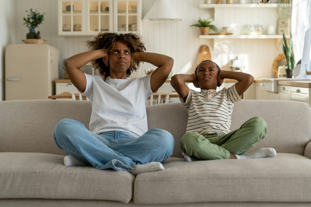 Annoyed mom with kid boy sit on sofa cover ears with hands look up suffer from noise from neighbours